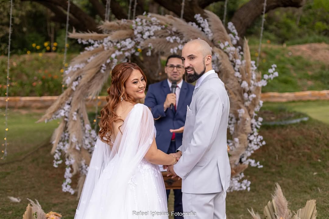 Noivos de mãos dadas sorrindo durante a cerimônia ao ar livre, com celebrante ao microfone e arco de flores secas ao fundo